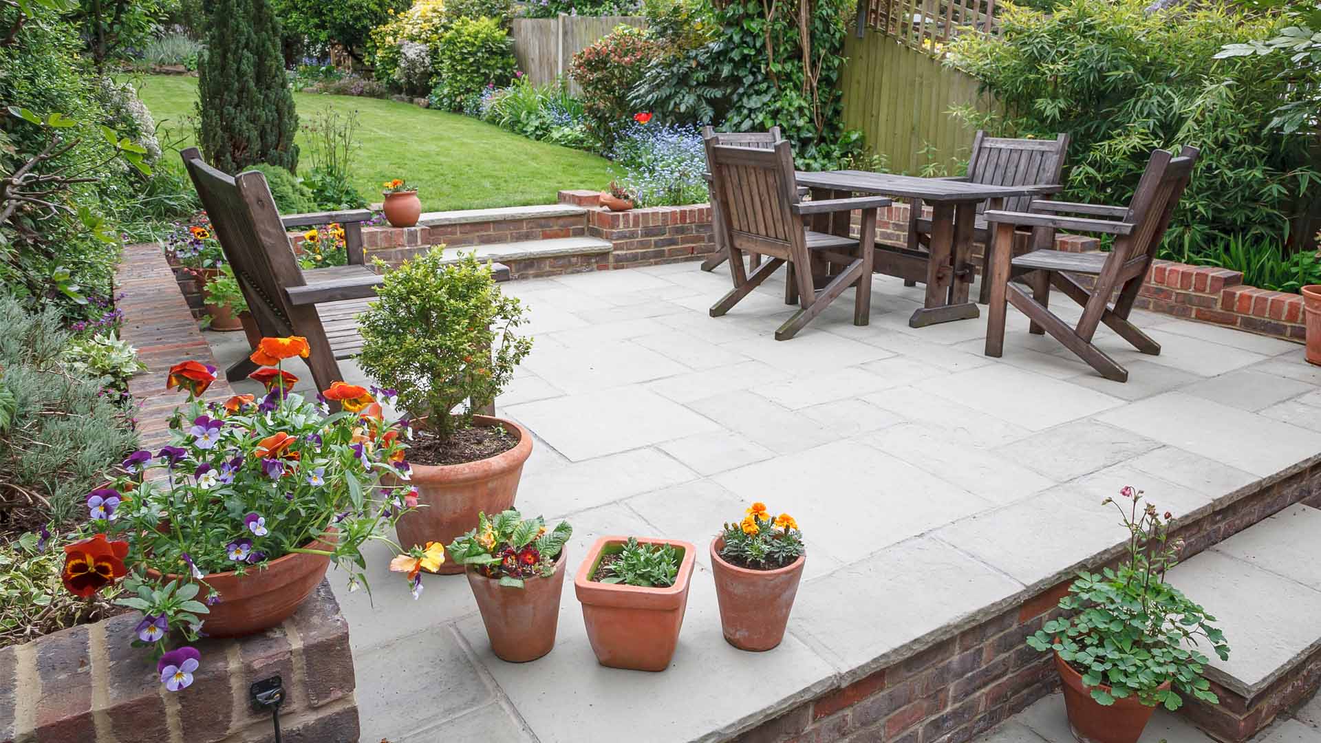 Brick Patio Installation with Lincs A garden patio with a stone surface features a wooden table and chairs. Potted flowers and plants, including vibrant orange and red blooms, line the edges, enriched by expert hydroseeding services in Western North Carolina. The backdrop reveals a lush green lawn with diverse shrubs and trees.