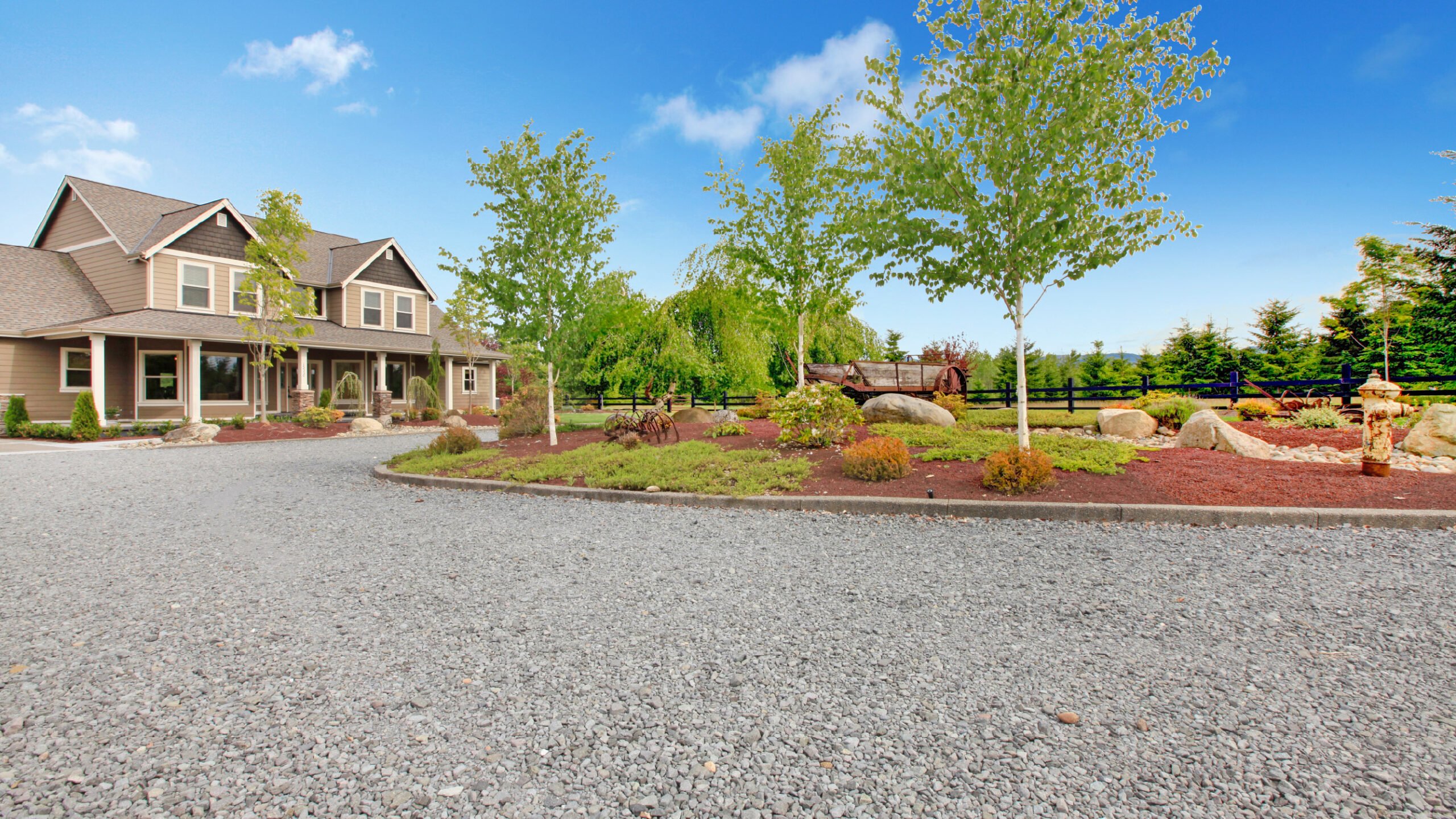 Gravel Driveway on Sunny Day A large, two-story house with a covered porch sits on a well-landscaped property. The yard features neatly arranged young trees, shrubs, rocks, and a newly refurbished gravel driveway leading to the house. If youre in Western North Carolina, consider local driveway repair services for top-notch upkeep.