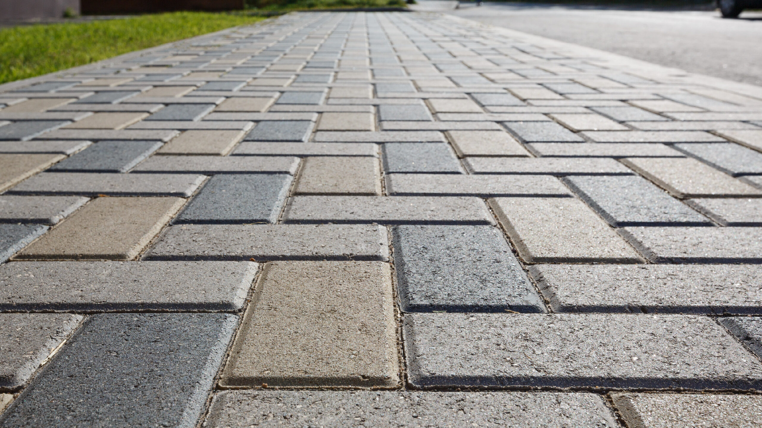 Paver Driveway with Lincs Close-up view of a paved walkway featuring a herringbone pattern with gray, beige, and light blue rectangular bricks. The sidewalk is bordered by lush green grass on the left and street on the right, offering inspiration for those considering driveway repair services in Western North Carolina.