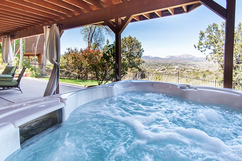 Bubbling hot tub under a covered patio with outdoor seating, overlooking a scenic mountain and valley landscape on a clear day.