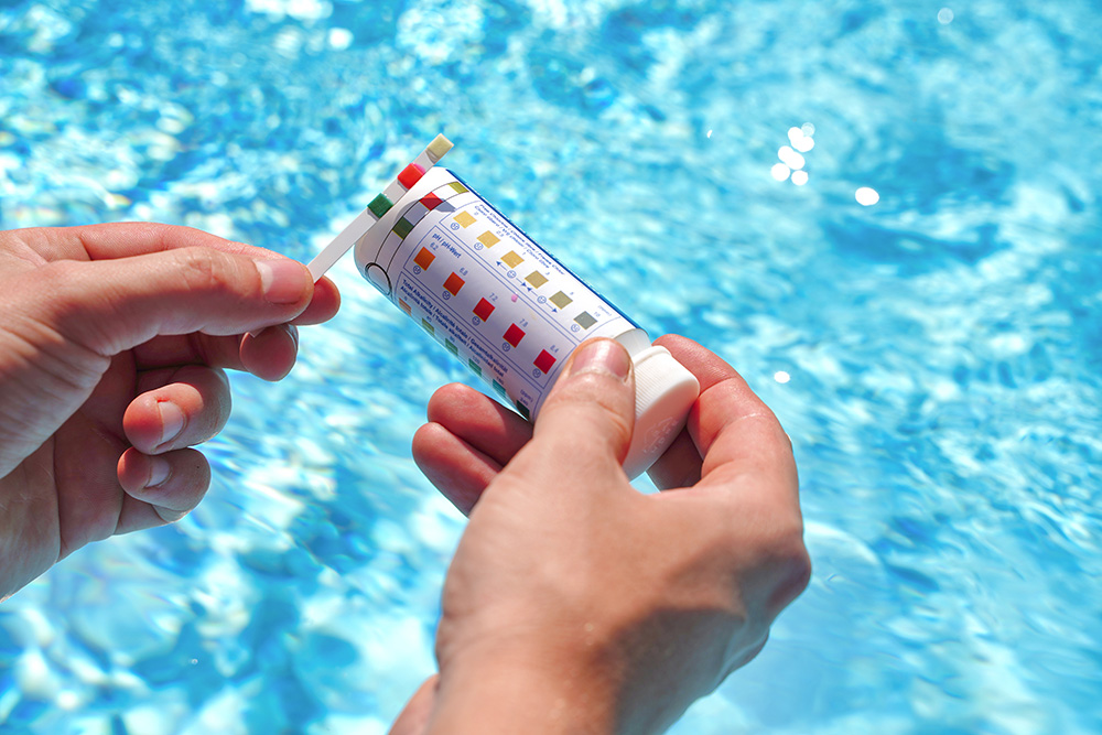A person holds a test strip and a container with a color chart, checking chemical levels in a swimming pool with clear blue water in the background.