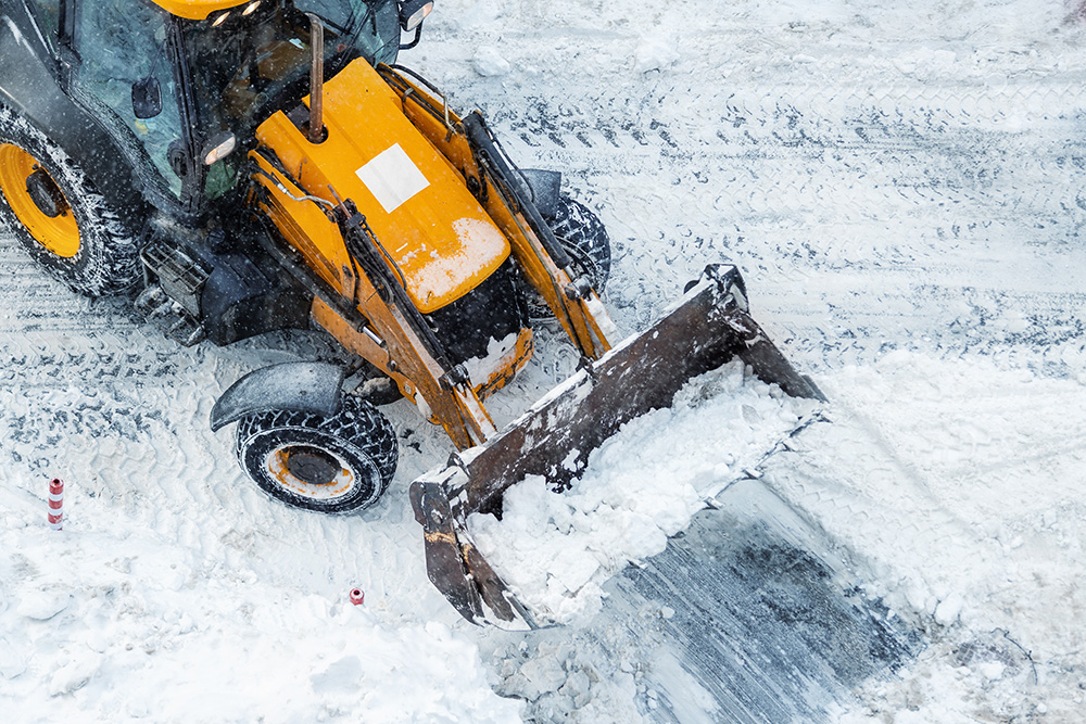 A yellow front loader clears snow from a road, with its bucket filled with snow, during winter. Snow covers the ground and vehicle, and tire tracks are visible on the icy surface.
