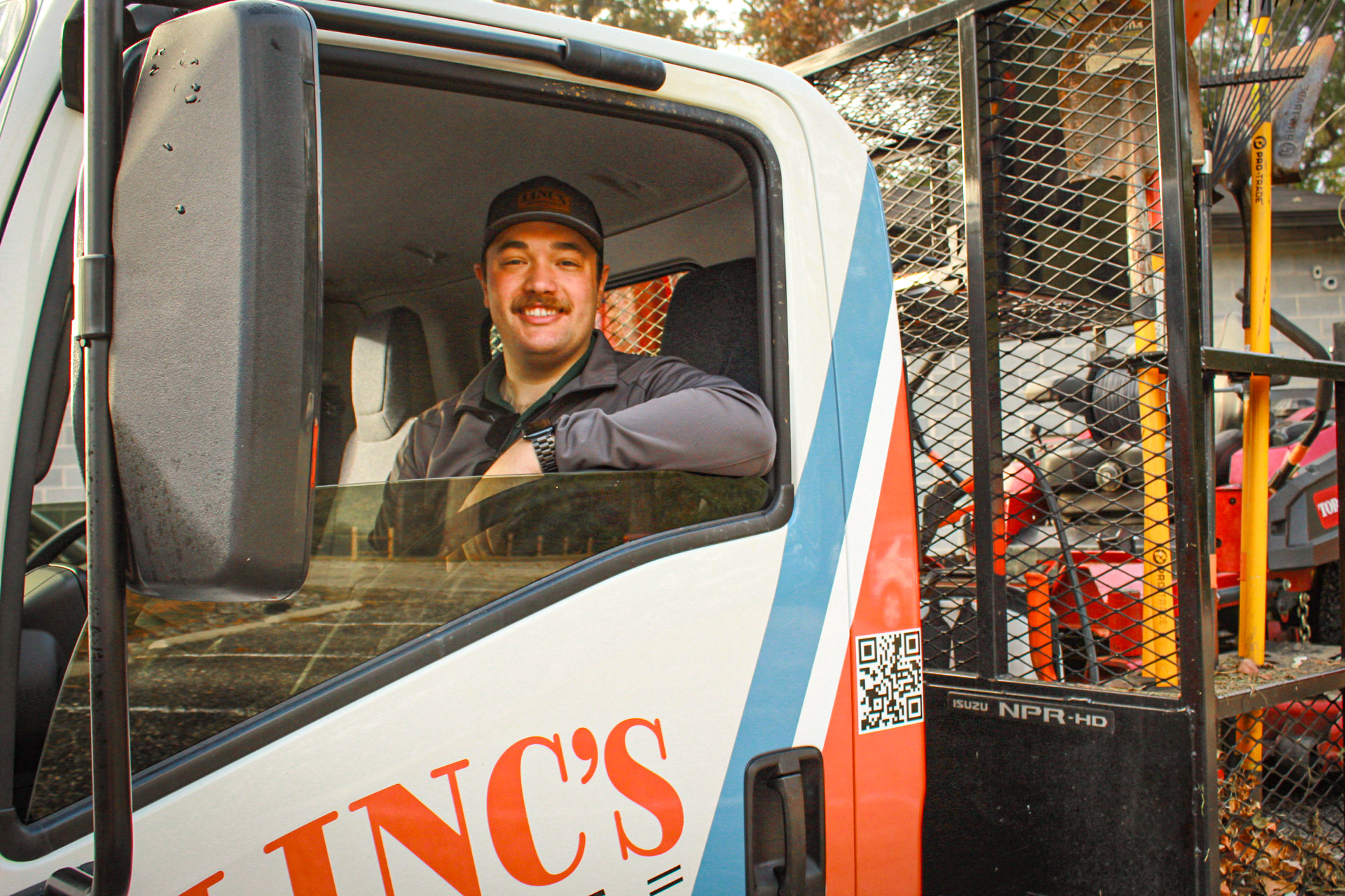 A man wearing a cap and jacket smiles while sitting in the driver’s seat of a truck with “INC’S” on the door, ready for year round maintenance. Equipment and tools are visible in the truck bed behind him.